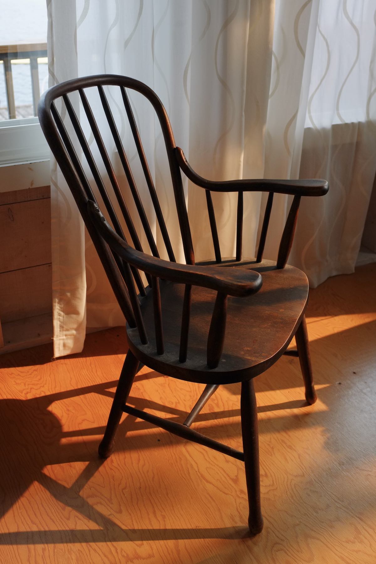 A dark wood armchair made in the Thonet bentwood technique, but more rustic. The heavy wooden seat is contoured as though to fit someone's butt. Late afternoon sunlight casts its shadow on the plywood foor. Translucent curtains are pulled partly shut behind it.