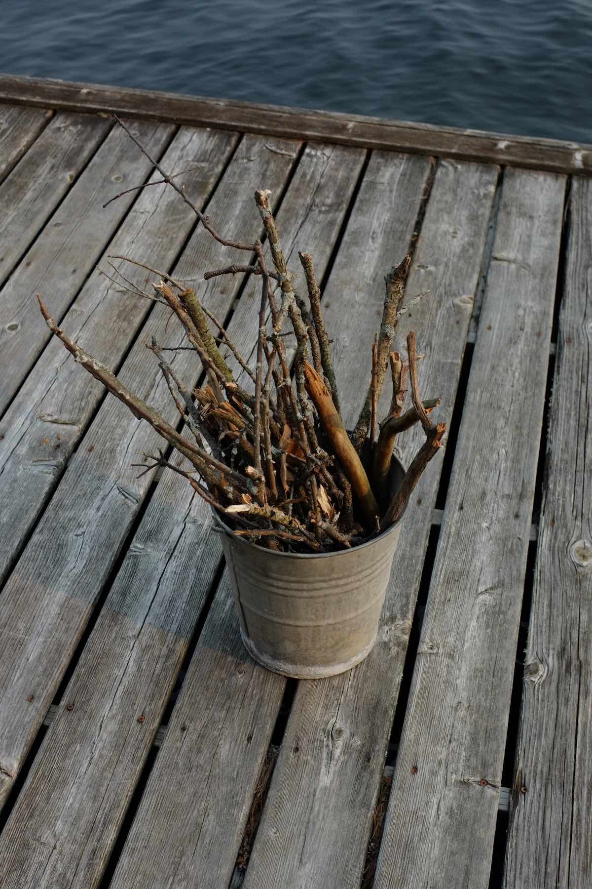 A weathered grey bucket with a variety of sticks sits on a wooden dock next to the water in the late afternoon sun.