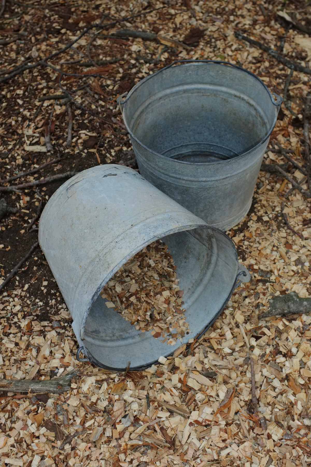 Two weathered grey buckets sit by a pile of woodchips, one on its side with a few woodchips spilling out.