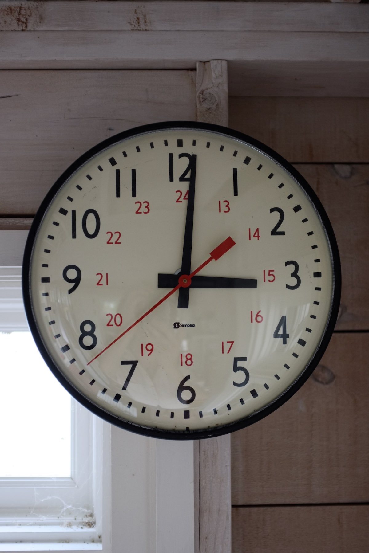 A clock showing three 3:00. It has simple straight hour and minute hands on a white dial with a red seconds hand. The numerals are set in Gill Sans, larger black numbers from 1–12 on the outside paired with smaller red numbers 13–24 on the inside.