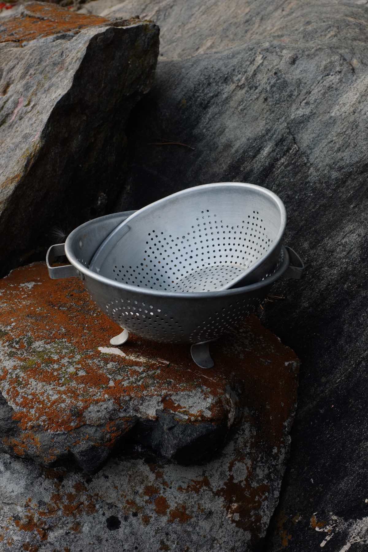 Two aluminum colanders sit on lichen-mottled rocks. The larger of the two has simple feet and handles.