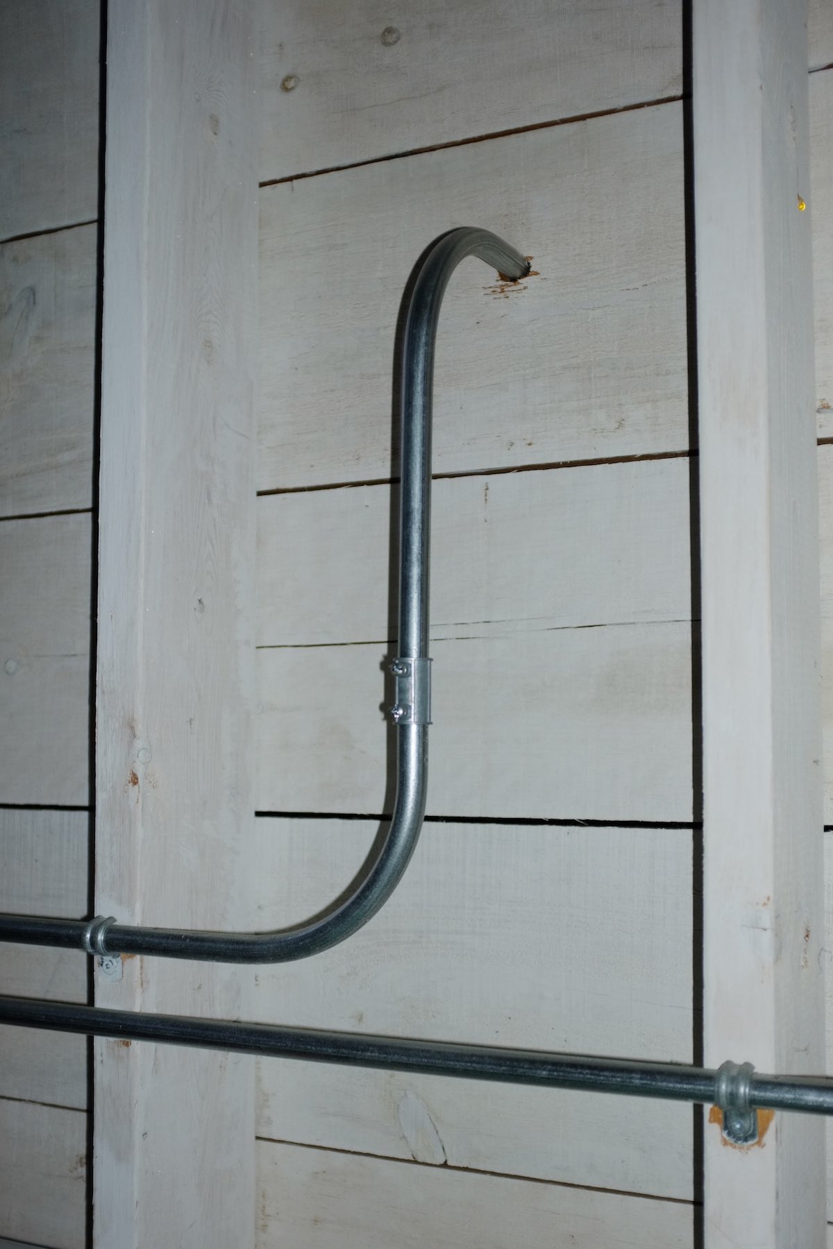 Metal electrical conduit elegantly curves up and into a hole in the white-stained wooden wall.