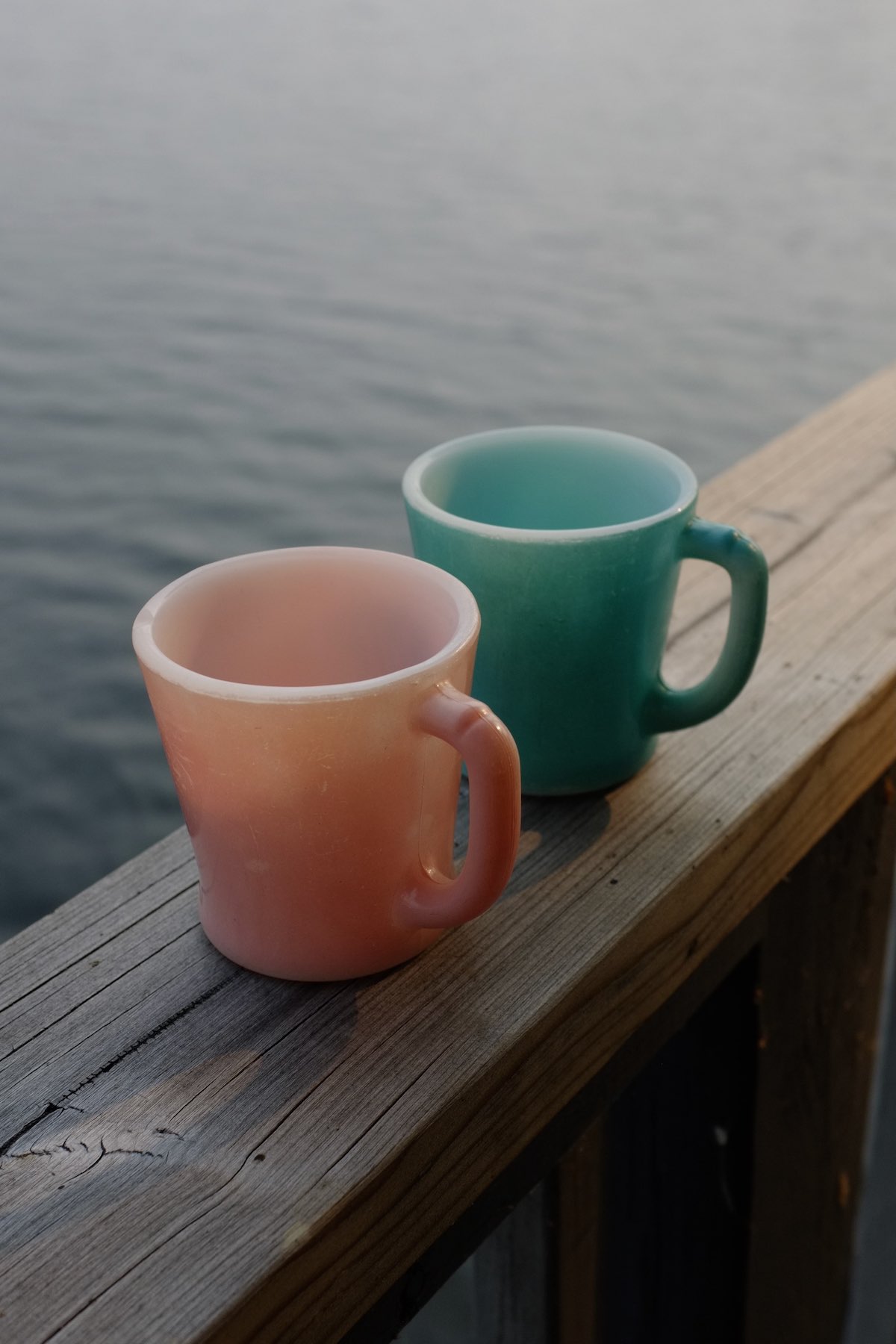 Two coloured glass mugs sit on a railing overlooking the water late in the day. They are light pink and dark turquoise, soft of a comfortable natural shape.