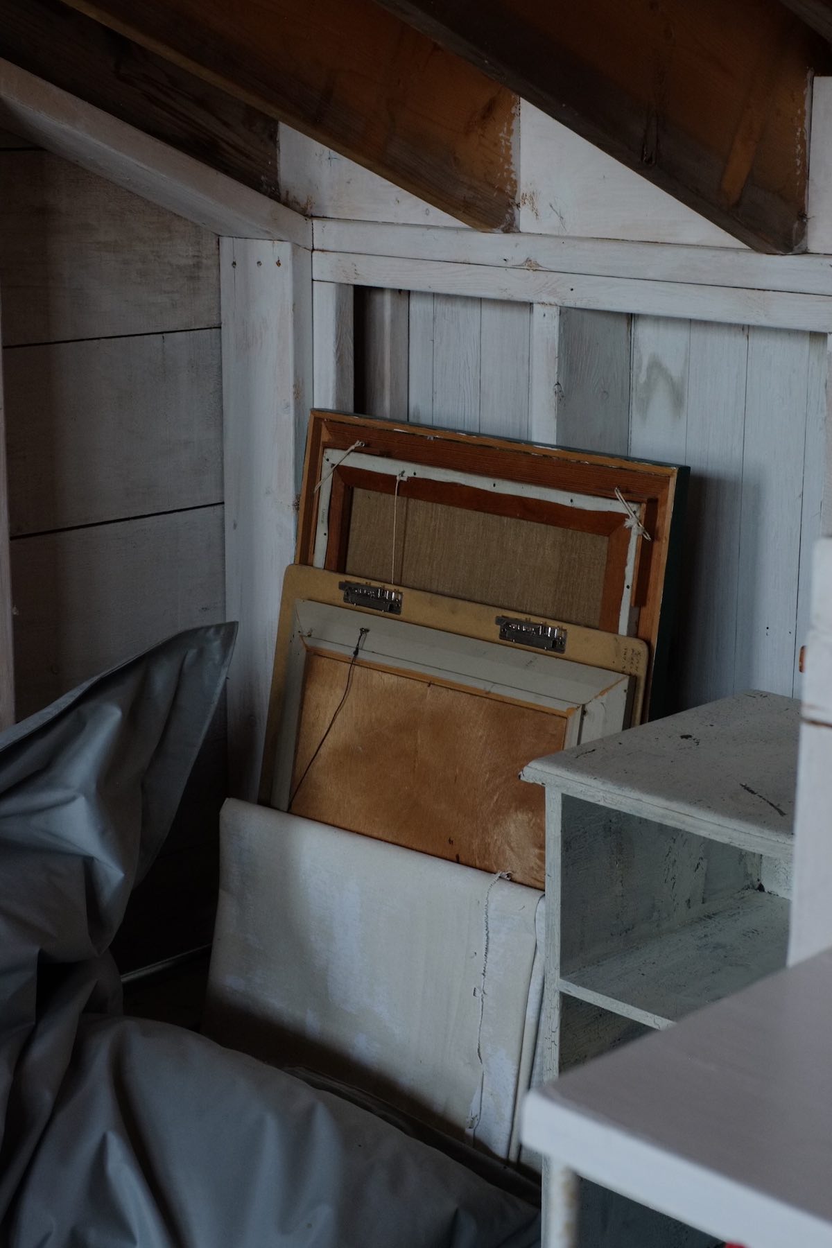 In a somewhat cluttered corner of a white-painted wooden room, a stack of old picture frames along with a folded white canvas with some white paint splatted on it.