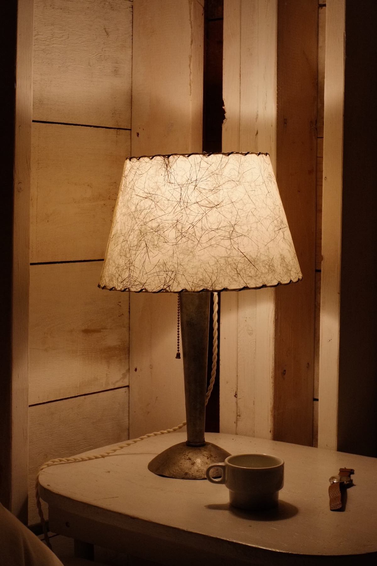 In a dim corner of a white-painted wooden room, an old hammered metal lamp with a lampshade containing scattered fibres glows softly. It sits on a small white table with a mug and a wristwatch.