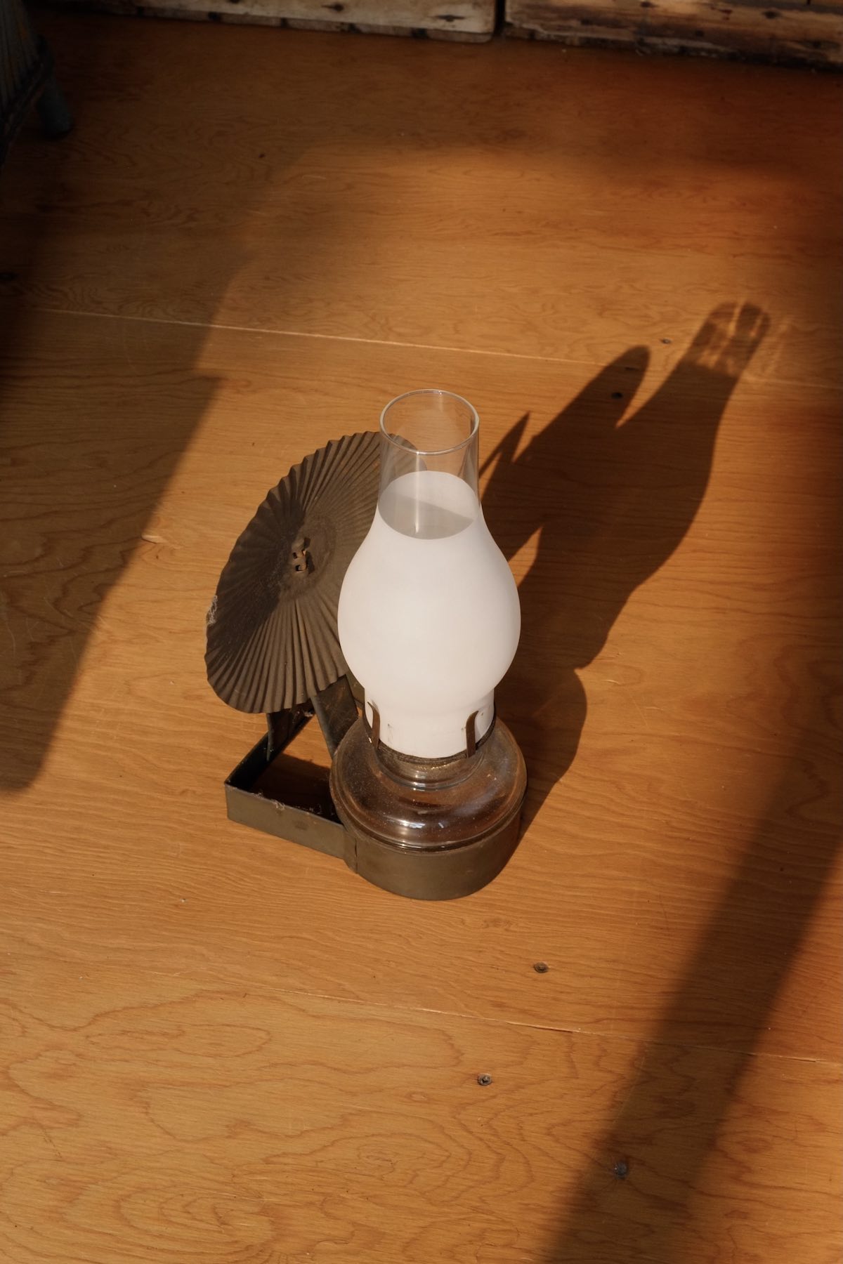 Glass oil lantern with a sheetmetal base and shade. The glass part smoothly flares out and tapers to a narrow top, mostly frosted white. The shade has radial ridges. It sits on a sun-drenched plywood floor.