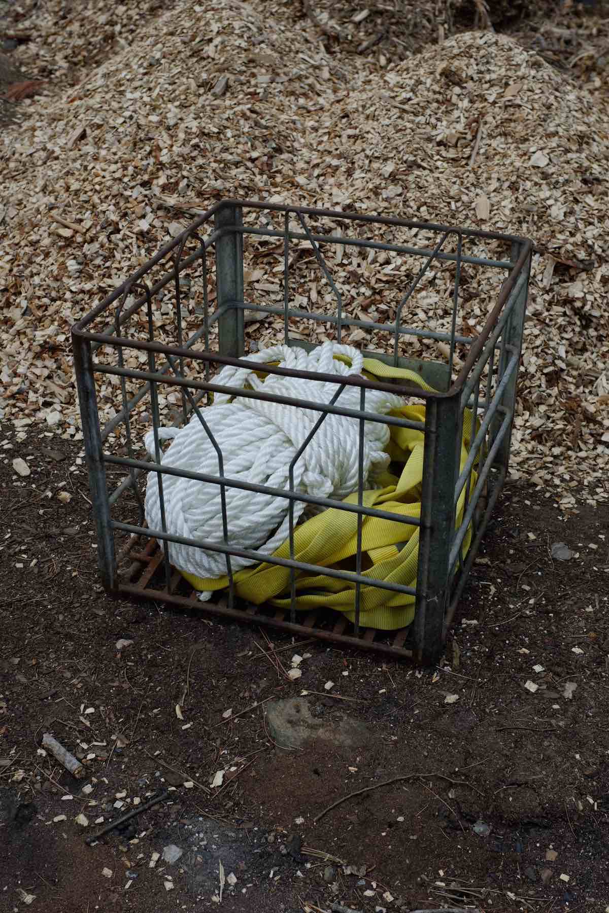 On the ground in front of a pile of woodchips sits an old rusting milk crate, a cubic structure of metal bars or tubes. Inside is a bundle of rope and a tie-down strap.