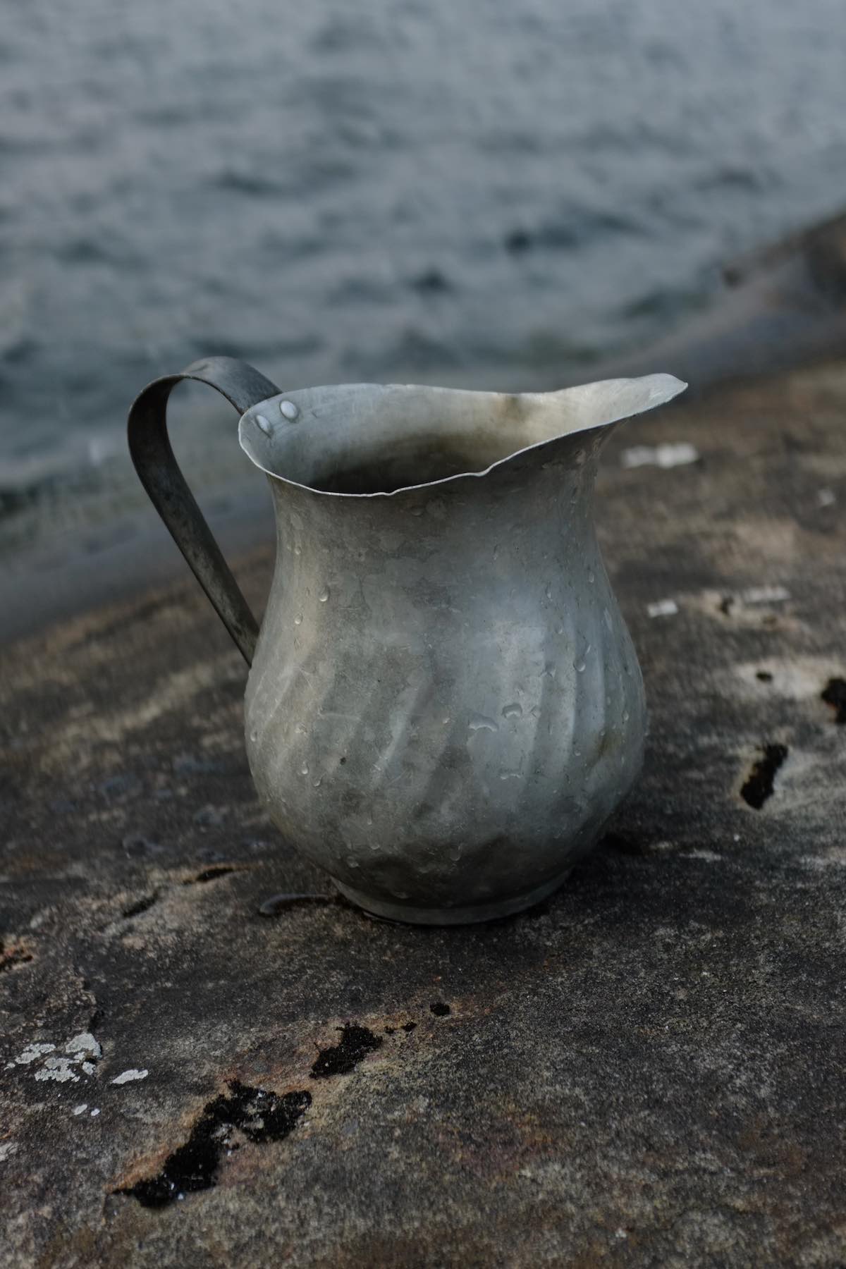 An aluminum pitcher sits on stone in front of the water. It has a large smoothly shaped spout and some texture on its lower section.