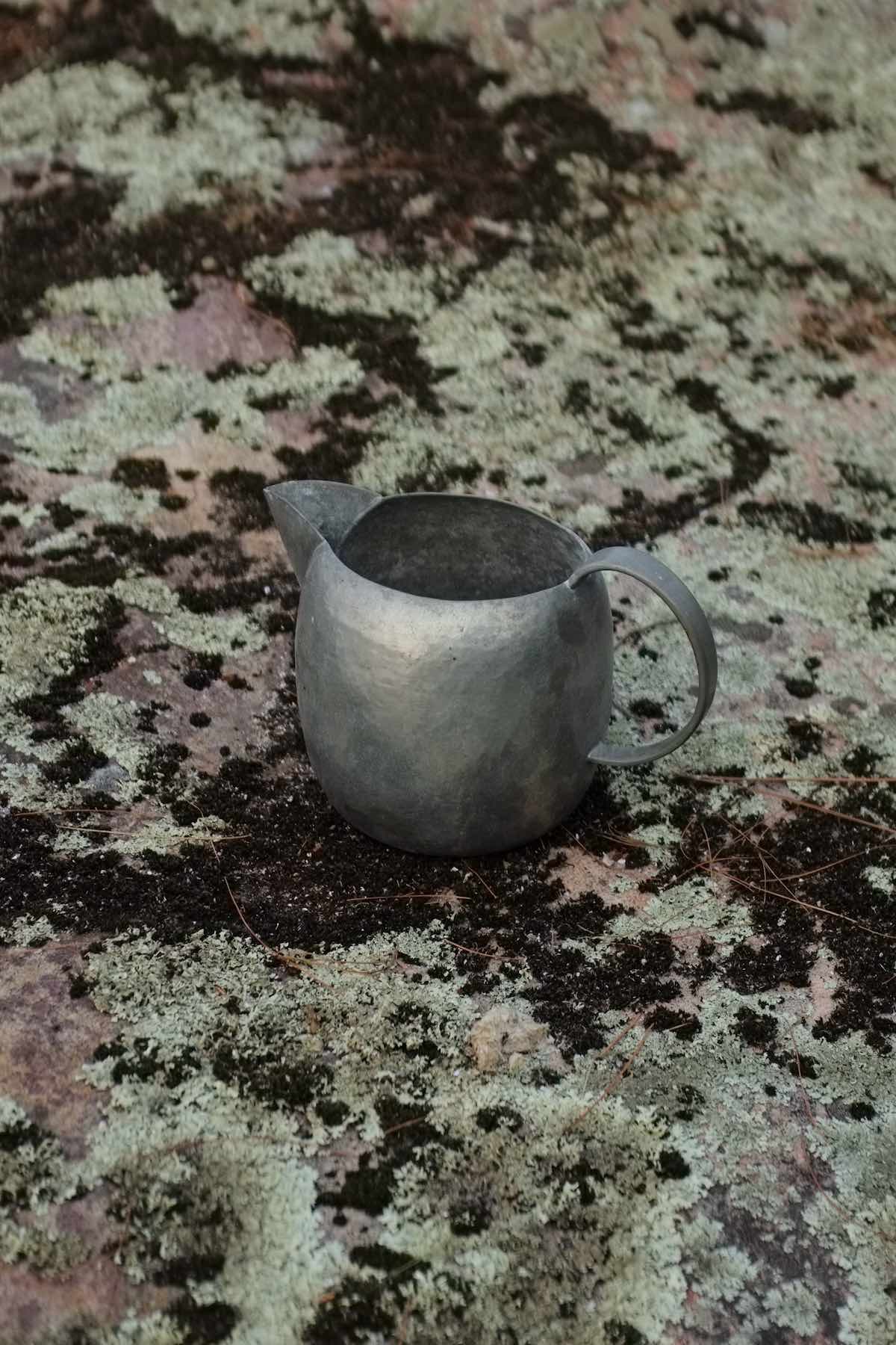 A more geometric pitcher — very round body and handle and a pointy spout. Hammered texture all over. It sits on lichen-covered natural pink granite.