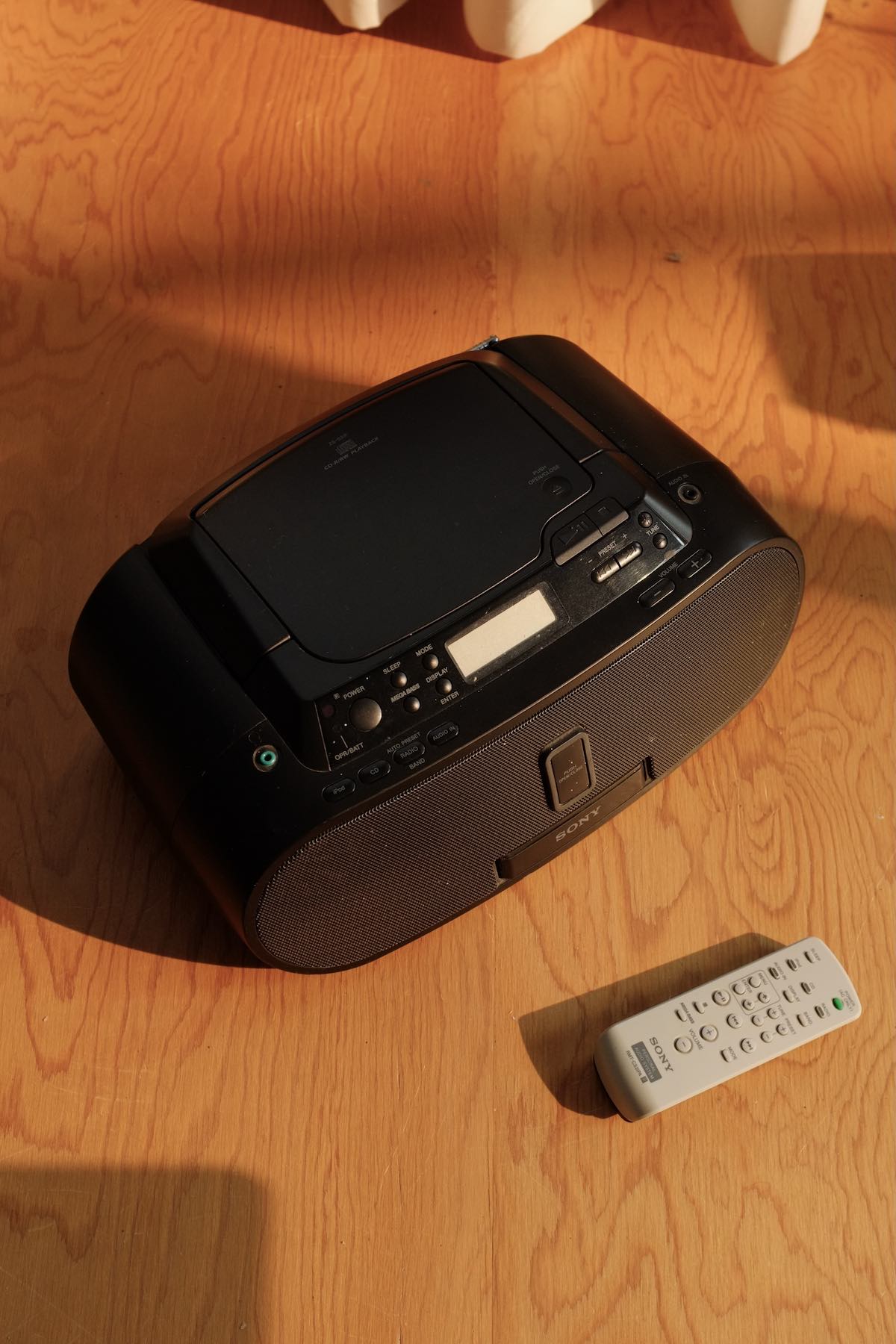2000s black Sony CD boombox with a light grey remote in the sun on a plywood floor. Generally amorphous design with a speaker grille covering the pill-shaped front.