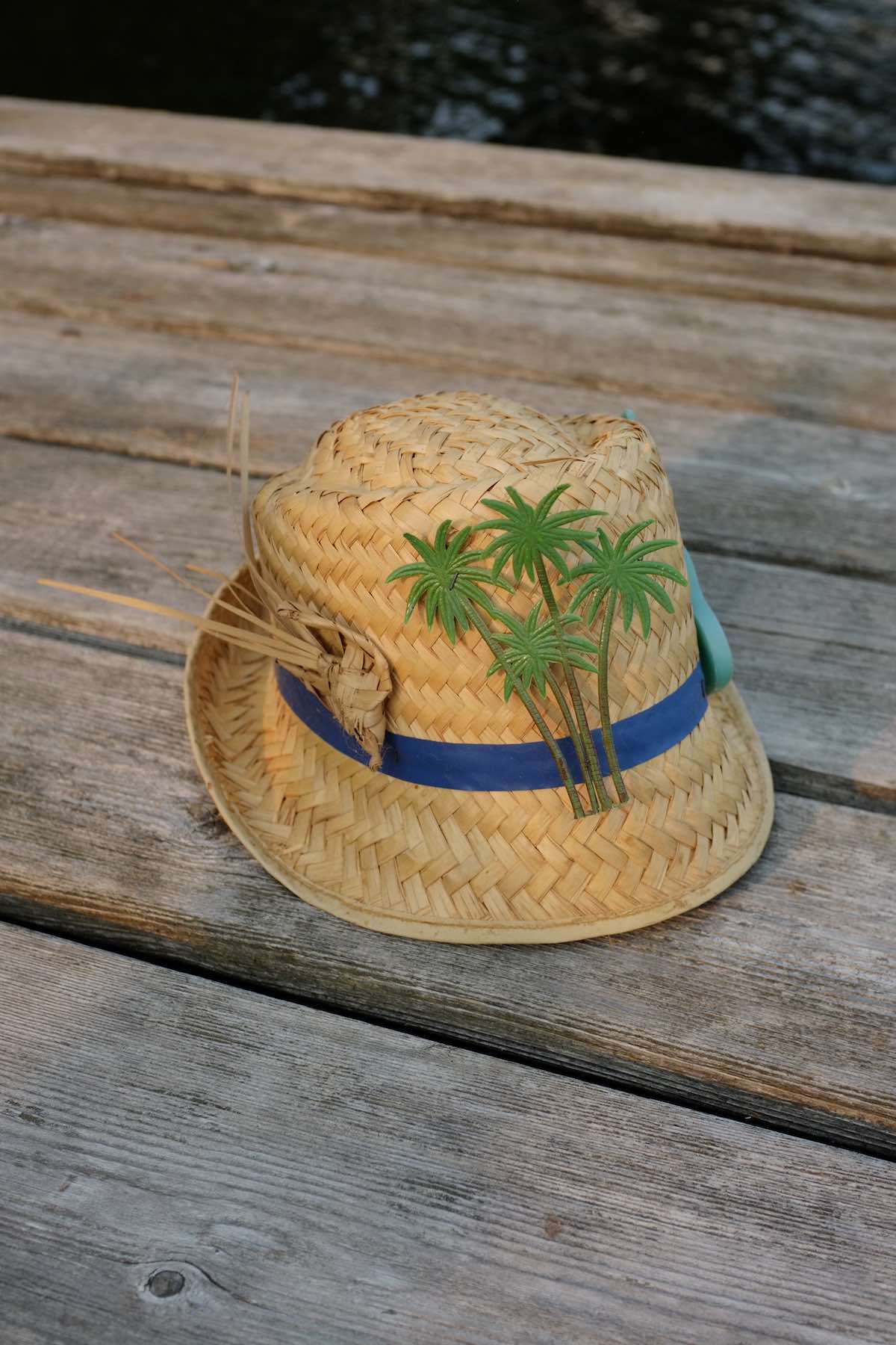 Small straw hat with a blue band around it. It has three decorations: an abstract bird made of the same straw, a plastic cluster of palm trees, and a green plastic acoustic guitar. It sits on a wooden dock in the late afternoon sun.