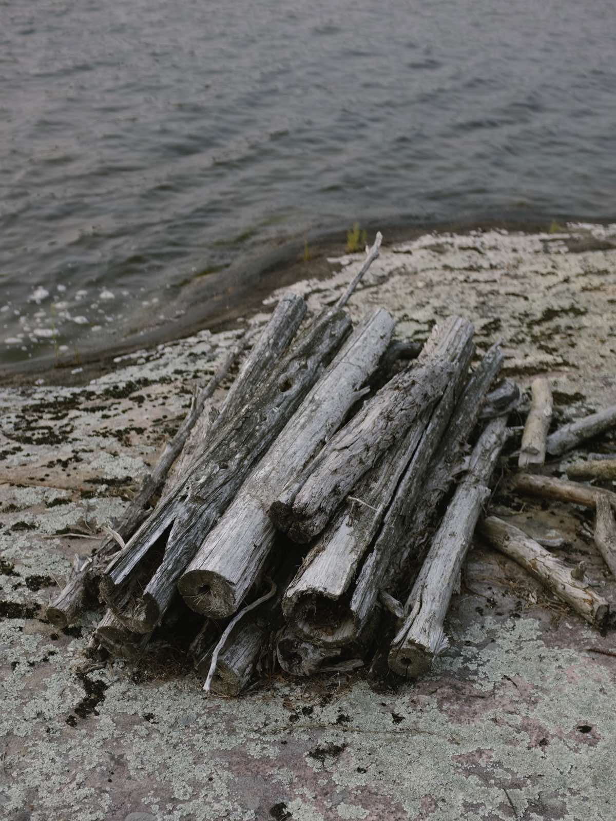 Logs from aforementioned construction set piece stacked like firewood on pink granite lichen-covered stone with lake water in background.