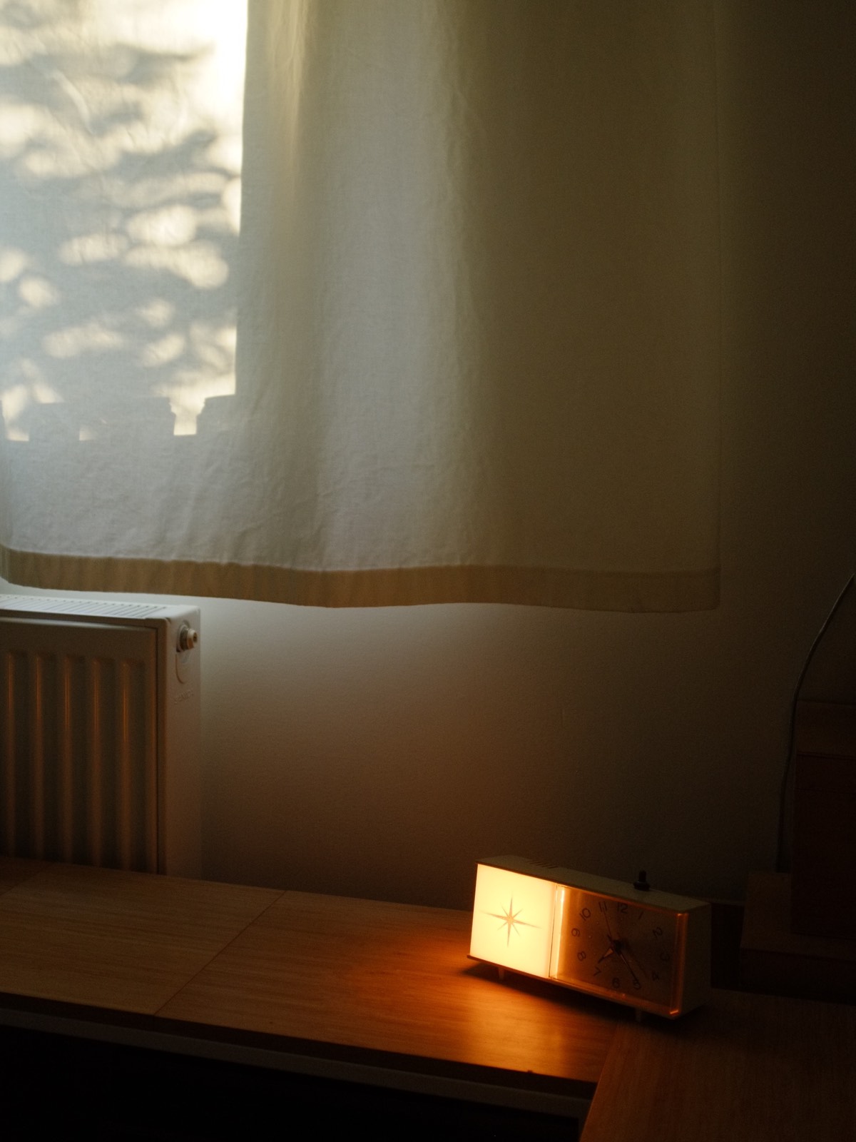 Glowing midcentury kitsch alarm clock on a sparsely decorated sideboard, early morning light coming through the curtains with tree branch shadows.